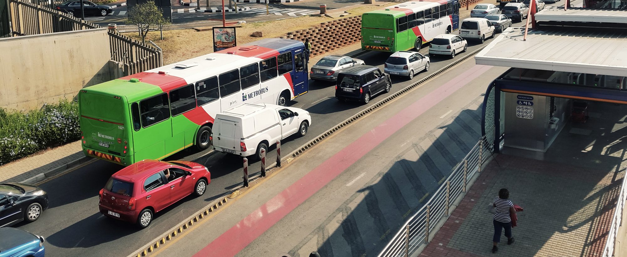 Two green and white Metrobus buses in mixed traffic beside the red Rea Vaya busway and a BRT station, seen from a footbridge
